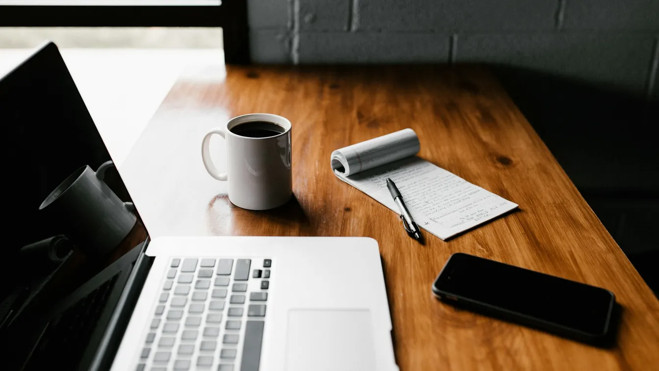 a laptop on a wooden desk with a notebook and a cup of coffee